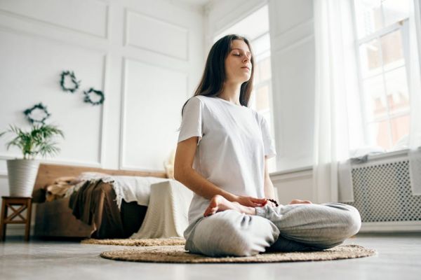 a young beautiful woman is sitting in a bright room on a jute rug and meditating with her eyes closed