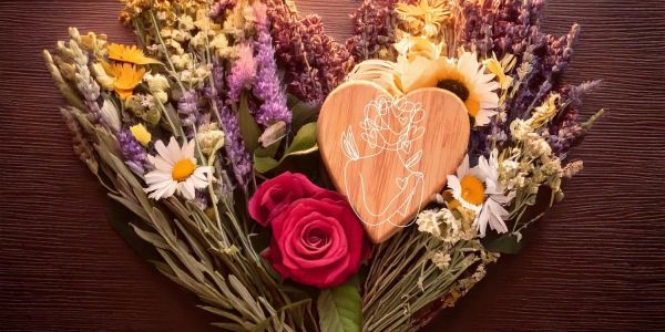 This beautiful image features a heart-shaped arrangement of vibrant herbs and flowers such as roses, lavender, chamomile, sage, and mugwort, centered with a wooden heart.