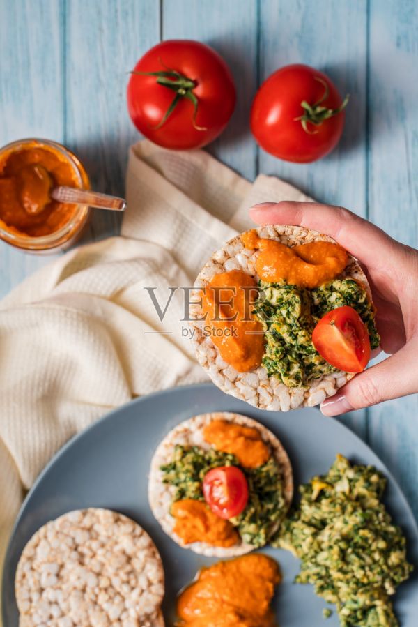 酥脆蓬松的年糕放在桌上，抹上鹰嘴豆泥、菠菜、鸡蛋、西红柿和蔬菜放在桌面上，手捧着健康的素食早餐无麸质-亮滤照片摄影图片