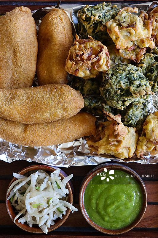 全幀圖像:木制托盤(pán)自制青椒油餅(mirch pakora)，酥脆的洋蔥bhajis，在錫紙上的palak pakora(菠菜油餅)，旁邊是一碗薄荷和香菜酸辣醬，磨碎的奶酪，印度美食素食小吃，從上方看圖片素材