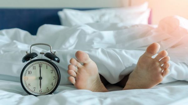 world sleep day, sleeping hour, health relaxation and bedtime insomnia concept with young girla??s bare foot laying on bed resting in bedroom on white bed sheet with alarm clock