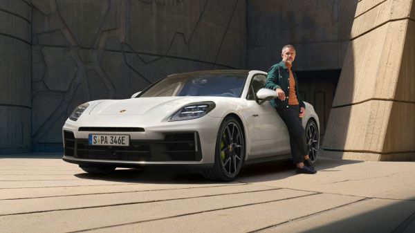 停放在混凝土墙前的细花白金属漆保时捷 Panamera 4S E-Hybrid 从前面看的前侧视图一名男子倚在驾驶员侧车门上
