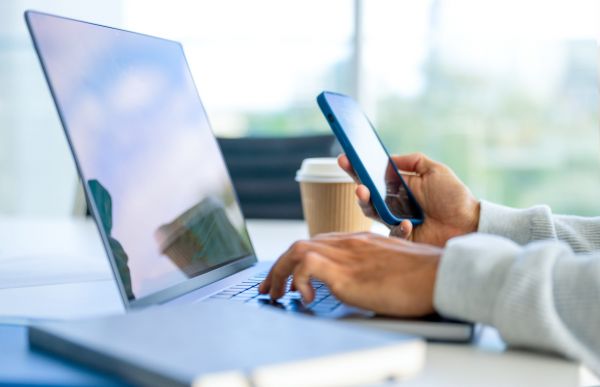 one hand typing on a laptop on a table and another hand holding a cell phone