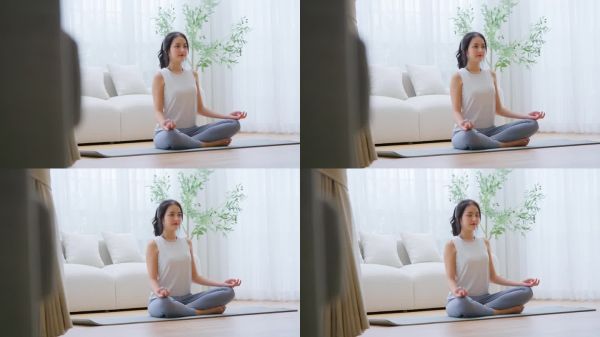 快乐的年轻亚洲女性在家中练习瑜伽和冥想，坐在客厅的地板上，双腿交叉，闭眼放松。正念冥想与身心健康的概