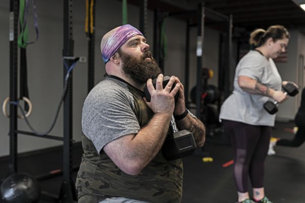 A full figured man and woman use dumbbells to do goblet squats while attending a cross training group fitness class at their local gym.
