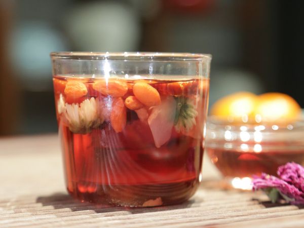 一位女性坐在窗邊書桌前，手持透明玻璃杯飲用玫瑰花枸杞茶，窗外陽光灑入，桌上放有筆記本和打開的茶葉罐，展現(xiàn)寧靜健康的生活方式