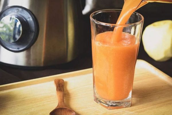 一杯透明玻璃杯中盛有淡琥珀色液體，杯壁可見細(xì)膩氣泡，旁邊放置一勺天然結(jié)晶蜂蜜和一小瓶手工蘋果醋，背景為木質(zhì)餐桌與新鮮蘋果