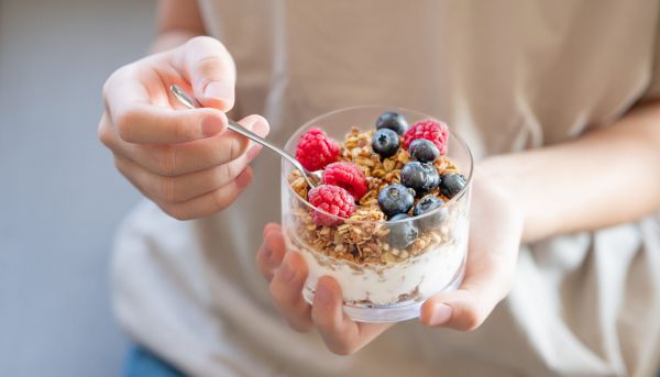 granola with greek yogurt, raspberries and blueberries in a glass in hands of a woman in a light t shirt close up. dairy breakfast concept, oat muesli in the morning light.