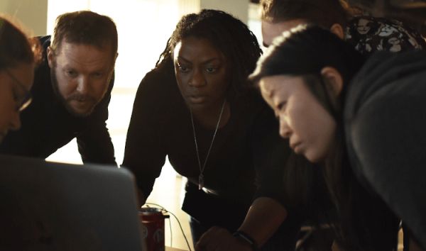 一群人，包括一名白人男子、一名黑人女子和一名亞裔女子，圍在一臺筆記本電腦旁。