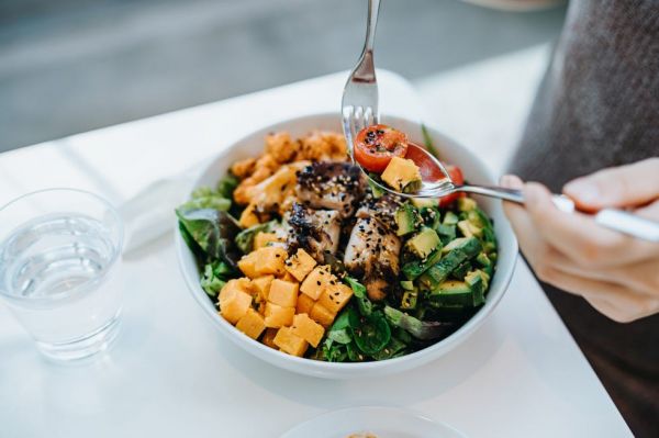 close up, high angle shot of young woman enjoying multi coloured healthy fruit, vegetables with grilled chicken salad bowl with balanced nutrition in cafe, with a glass of water by the side healthy eating lifestyle people, food and lifestyle concept